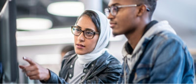 Dwie osoby pracujące razem przy komputerze. Kobieta w białej chuście na głowie wskazuje na ekran, obie osoby są skupione i zajęte rozmową.