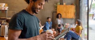 Smiling man holding a coffee cup and using a tablet in a cozy café, with a group of people chatting in the background.