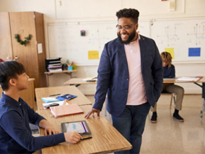 A teacher in a blue blazer smiles while conversing with a seated student in a classroom with a whiteboard in the background.