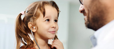  A young girl with a playful smile holds a stethoscope to her ears, looking up at an adult male in glasses.