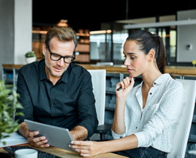 A man and a woman are sitting at a desk in an office, looking at a tablet together