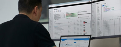 Person vid skrivbordet med tre bildskärmar som visar hotanalys, kontoinloggningar och meddelandeapp.