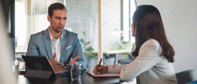 Twee professionals in een vergadering, een man die praat en gebaart terwijl een vrouw luistert en notities maakt.