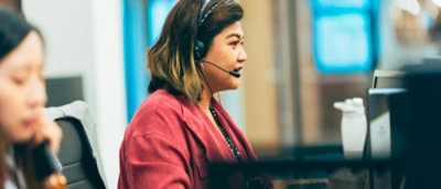Person in a red sweater seated at a desk in an office setting with computer monitors and another individual partially visible in the background.