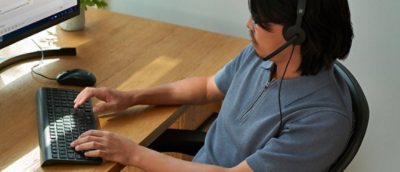 Person in a blue shirt typing on a keyboard at a wooden desk with a monitor, mouse, and coffee cup in a tech-focused workspace.