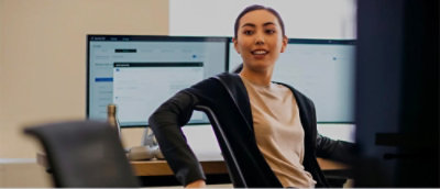 Individual in a light shirt and dark cardigan seated in an office chair with two monitors displaying web content behind them.