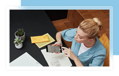 Eine Frau in einem blauem Hemd sitzt an einem Tisch mit einem Notebook, einem gelbem Umschlag und Topfpflanzen und benutzt ein Smartphone.