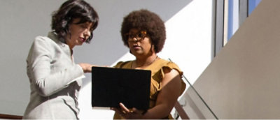 A woman wiaTwo women stand on a staircase, focused on a laptopth glasses works on a laptop at a desk, with a plant and window in the background.