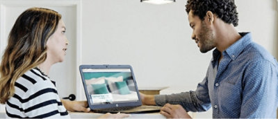 Two people are looking at a laptop screen displaying an image of a bed with pillows in a room setting.