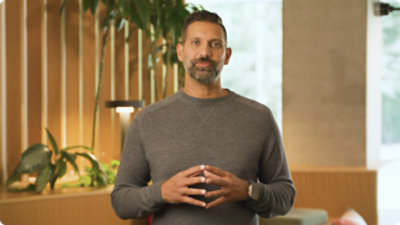 Un hombre con barba en un cuadro gris se encuentra en un entorno de oficina con plantas y una partición de madera en el fondo.