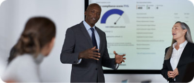 Un hombre con un traje presenta datos en una pantalla, mientras una mujer se encuentra junto a él, en un entorno de reunión profesional.