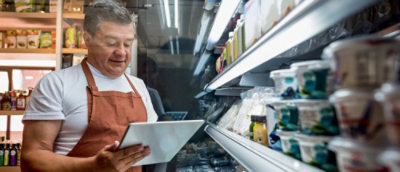 Ein Mann in einer orangefarbenen Schürze, der ein Tablet hält, vor einem Regal mit Lebensmitteln