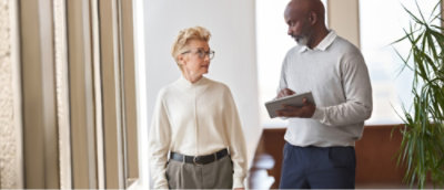 Dwie osoby stoją w pomieszczeniu, jedna z nich trzyma tablet. Wyglądają na pogrążone w rozmowie. W pobliżu widać roślinę.