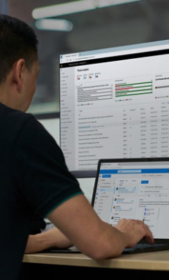 En person sitter vid ett skrivbord och arbetar på en bärbar dator med en stor datorskärm framför som visar olika systemdata
