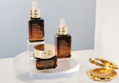 Three Estée Lauder Night Repair bottles, a gold jar, and powder compact on a clear stand in an elegant, luxe setting.