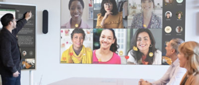 Una reunión empresarial con un hombre presentando a compañeros en una sala y otros que participan virtualmente en una pantalla grande.