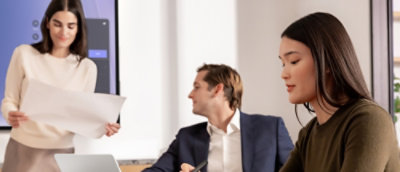 Grupa ludzi siedzi przy stole. Obok stoi kobieta trzymająca kartkę papieru, a obok mężczyzna w garniturze.