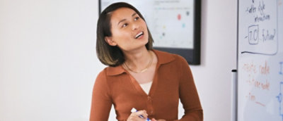 A woman in a brown shirt standing in front of a whiteboard with writing.