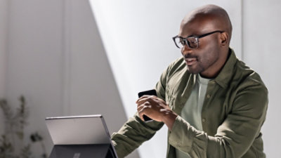 A man with glasses smiles at a tablet while holding a smartphone and wearing a green jacket.