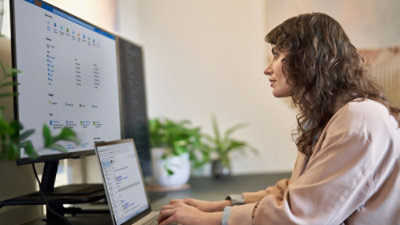 A woman sitting at a desk looking at a laptop and a large monitor screen.