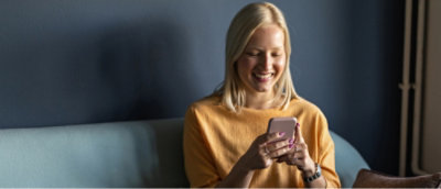 Una mujer con un suéter amarillo sonriendo mientras mira su smartphone, sentada en un sofá azul.