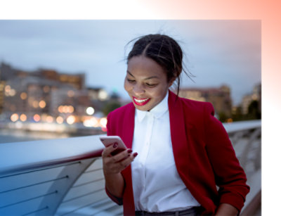 A smiling woman in a red blazer using a smartphone on a city bridge at dusk.