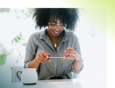 A smiling woman in glasses, using a smartphone at a table with a coffee mug beside her.