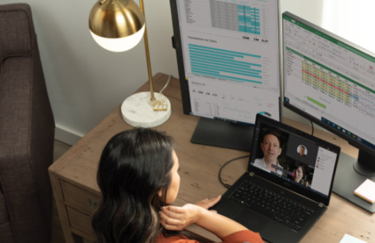 Osoba siedząca przy biurku i uczestnicząca w połączeniu wideo w usłudze Teams na laptopie przed dwoma monitorami stacjonarnymi.