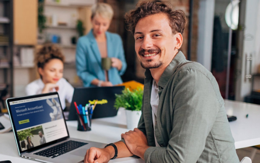 A cheerful man in front of a laptop sits at a conference table across from his two female colleagues.