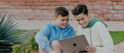 Två pojkar i huvtröjor sitter på gräset och ler medan de tittar på en öppen bärbar dator.