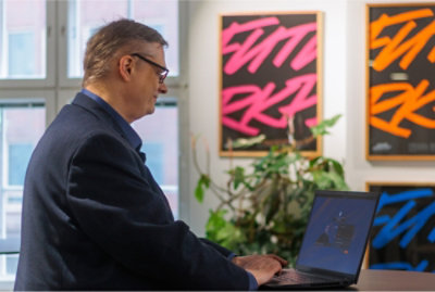 Osoba w garniturze pracuje na laptopie w biurze z kolorowymi grafikami i roślinami w tle.