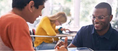 Dwie uśmiechnięte osoby siedzące przy stole i rozmawiające ze sobą. W tle widać inną osobę skupioną na tablecie.