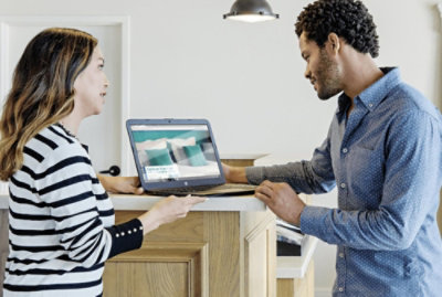 Two people stand at a counter looking at a laptop. One person is holding the laptop and speaking