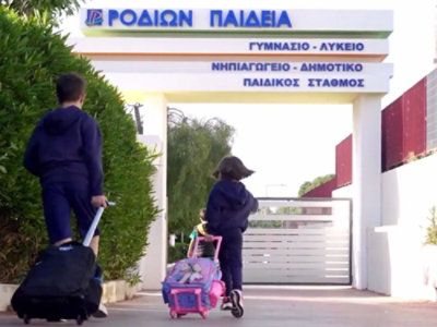 Dwoje dzieci z plecakami na kółkach idących w stronę wejścia do szkoły z napisem w języku greckim.
