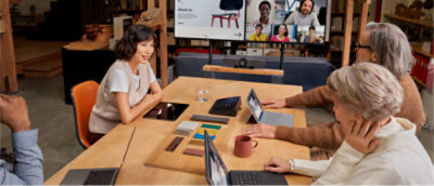 คนสามคนนั่งอยู่รอบโต๊ะพร้อมแล็ปท็อปและมีการประชุมทางวิดีโอพร้อมผู้เข้าร่วมหลายคนซึ่งแสดงบนหน้าจอขนาดใหญ่